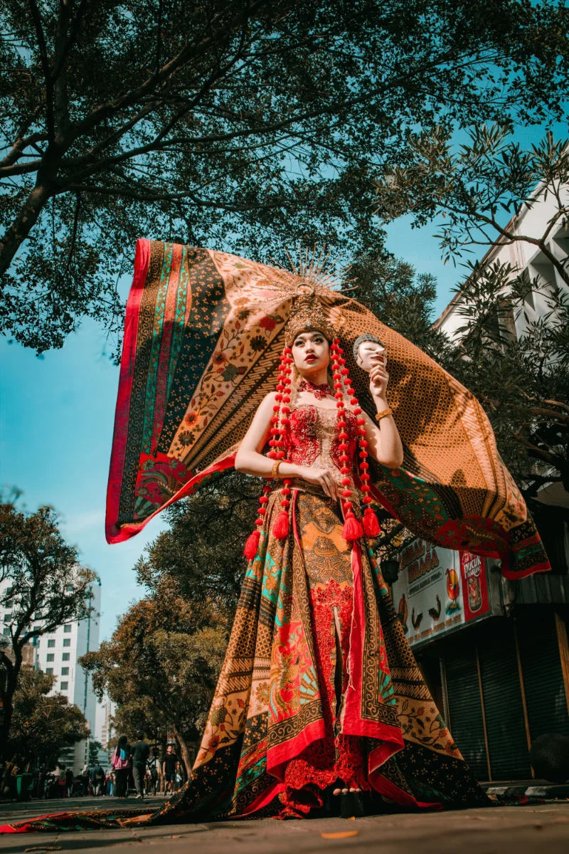 Model in a flowing batik-patterned gown with an ornate gold headdress and cascading red tassels, holding a white mask, standing on Braga street.